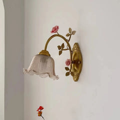 Close-up view of a gold floral wall sconce featuring a textured glass shade and pink rose accents, mounted on a white wall.