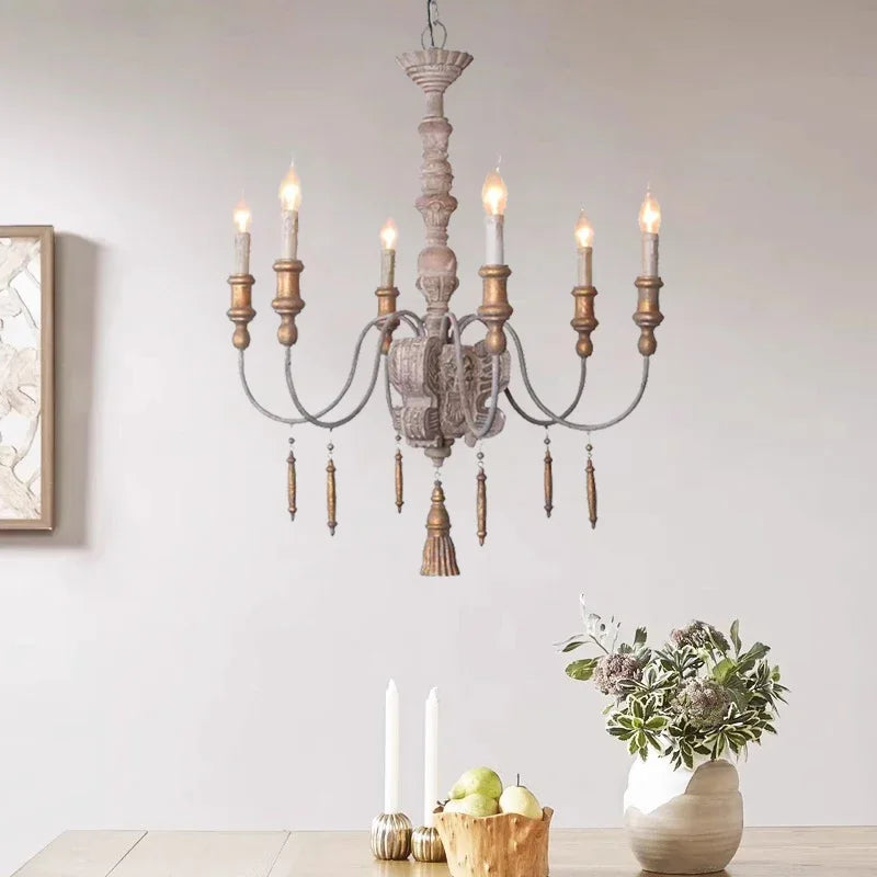 A distressed wood chandelier with candle-style lights and hanging tassels, illuminated and hanging in a room with a table setting.