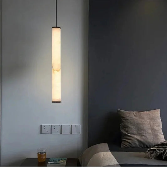 A long, slender marble cylinder pendant light with natural stone inclusions hanging in a dimly lit room next to a bed.