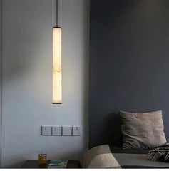 A long, slender marble cylinder pendant light with natural stone inclusions hanging in a dimly lit room next to a bed.