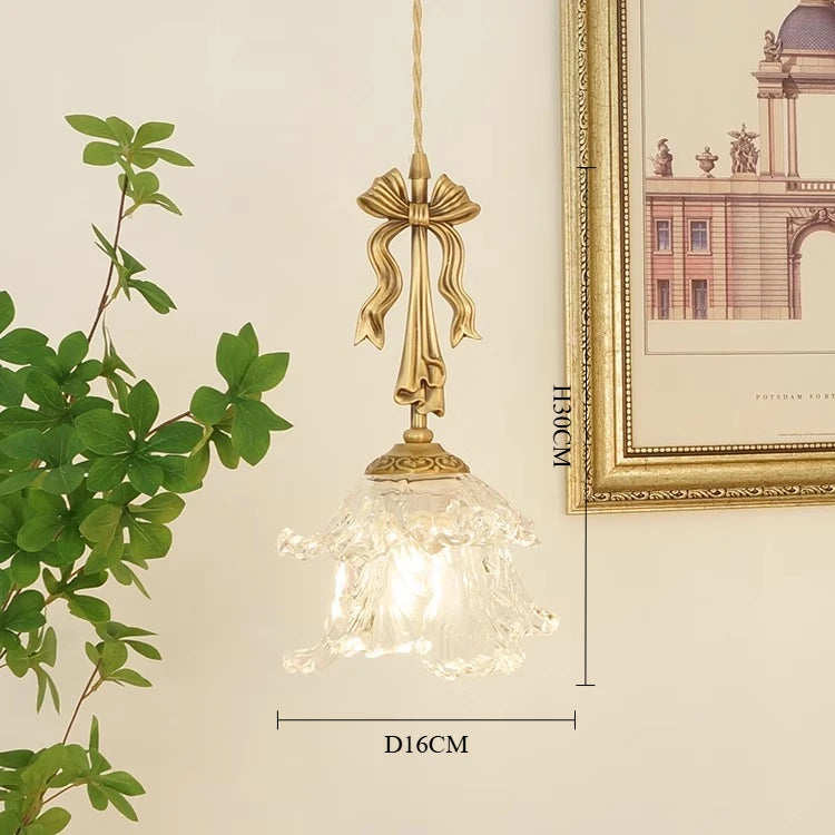 Decorative pendant light with gold bow design on a neutral wall with a plant and framed picture.