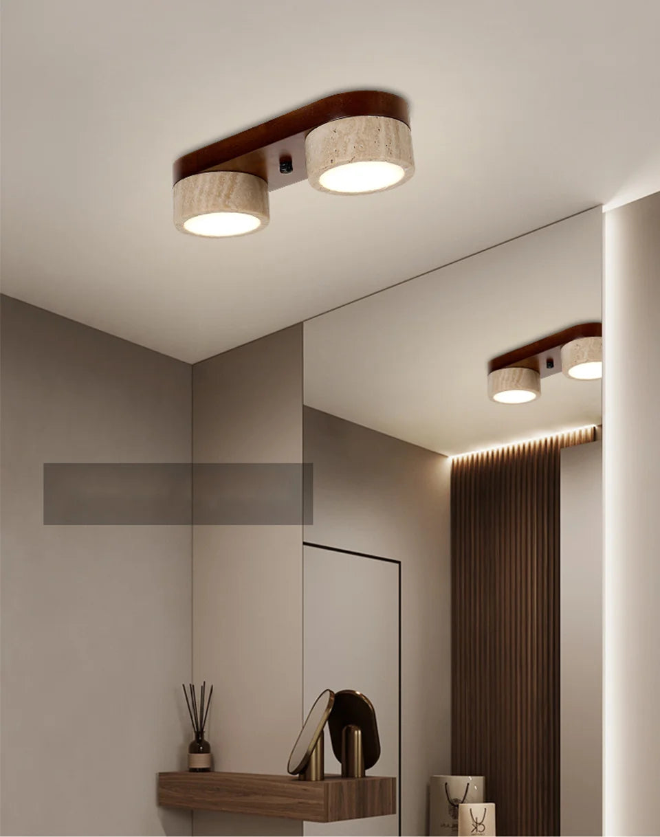 A hallway or bathroom with a modern flush mount ceiling light featuring two travertine stone cylinders on a dark wood base, reflected in a large mirror.