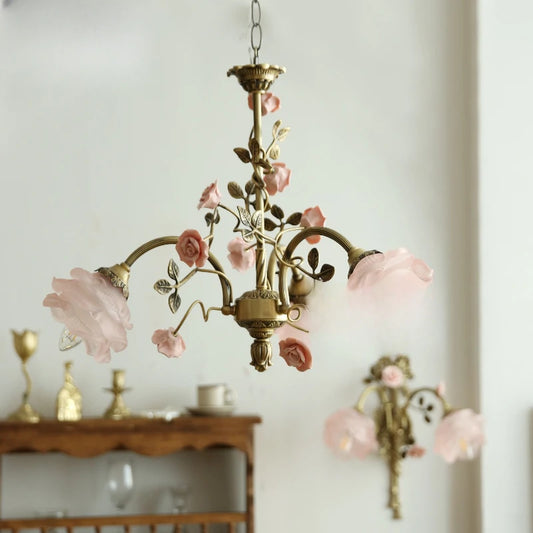 A brass chandelier adorned with pink rose buds and ceramic flowers, hanging against a white wall.