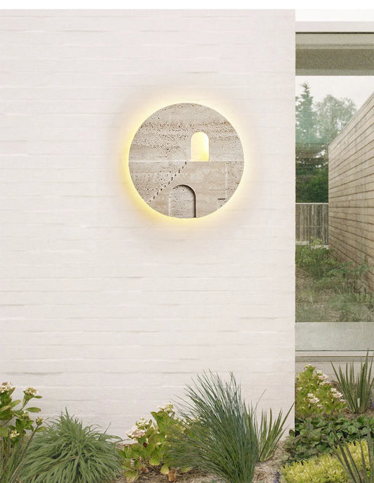 Round natural stone wall sconce with carved staircase and archway, mounted on a white brick wall outdoors.