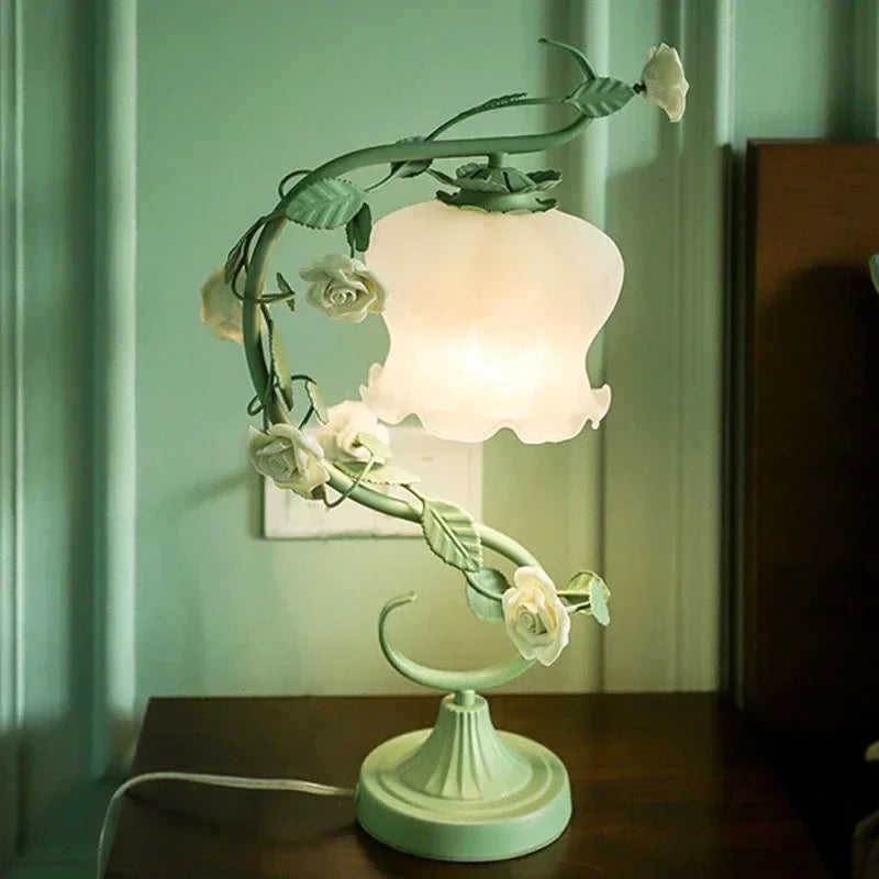 A green table lamp with a floral rose vine design and a white frosted bell-shaped shade, illuminated on a wooden surface.