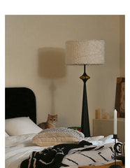 A dark grey floor lamp with a floral drum shade is positioned in a bedroom setting, casting a warm light.