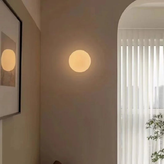 A warm glowing round glass wall light mounted on a beige wall in a room with sheer curtains.