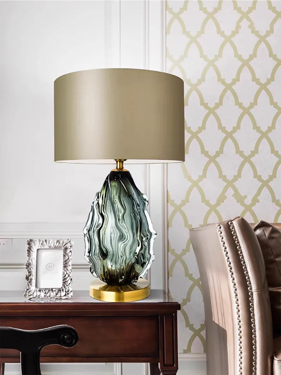 Decorative table lamp with a green and gold base on a wooden side table against a patterned wall.