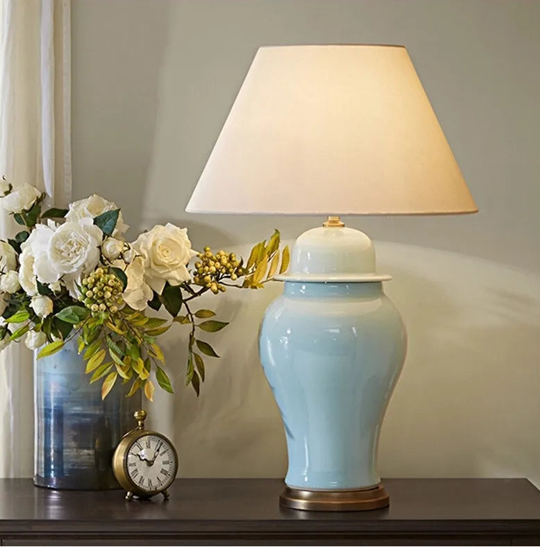 Close-up of a light blue ceramic table lamp with a cream drum shade, a brass base, and a small decorative clock.