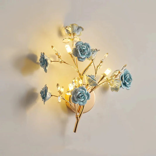 A gold wall sconce with blue ceramic flowers and clear crystals, emitting a warm light.