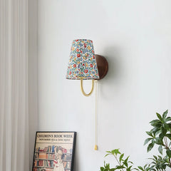 Wall-mounted lamp with floral patterned shade on a white wall with a plant and framed picture below. Accent wall lamp with floral lampshade and up-down LED lighting.