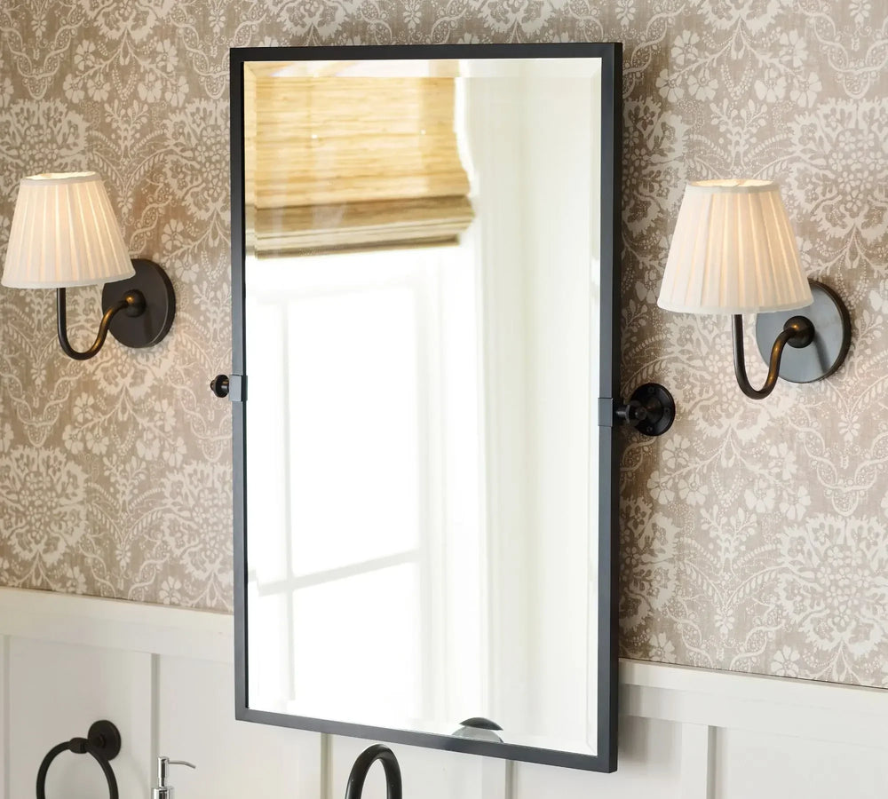 Rectangular mirror with black frame on a wall with decorative wallpaper and sconces.