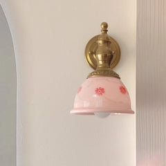 Wall-mounted light fixture with pink glass shade and gold metal fixture on a neutral wall.