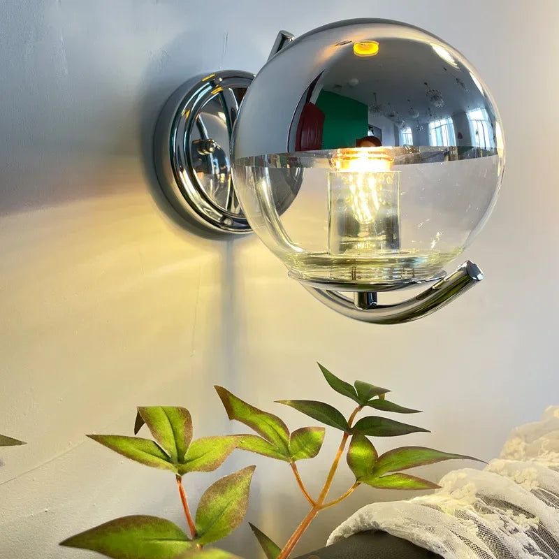 Wall-mounted light fixture with clear glass shade and metallic finish, illuminated against a neutral background.