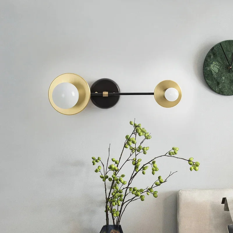 Modern wall lamp with gold and black design on a light gray wall with a plant and chair in the foreground.