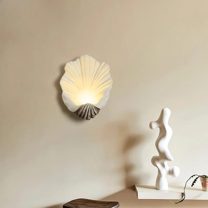 Shell-shaped wall light fixture on a beige wall with a sculpture and plant in the foreground.