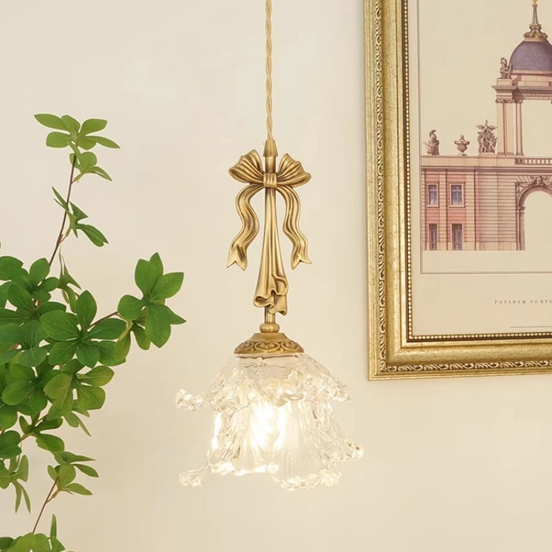 Decorative pendant light with gold bow design on a neutral wall with a plant and framed picture.