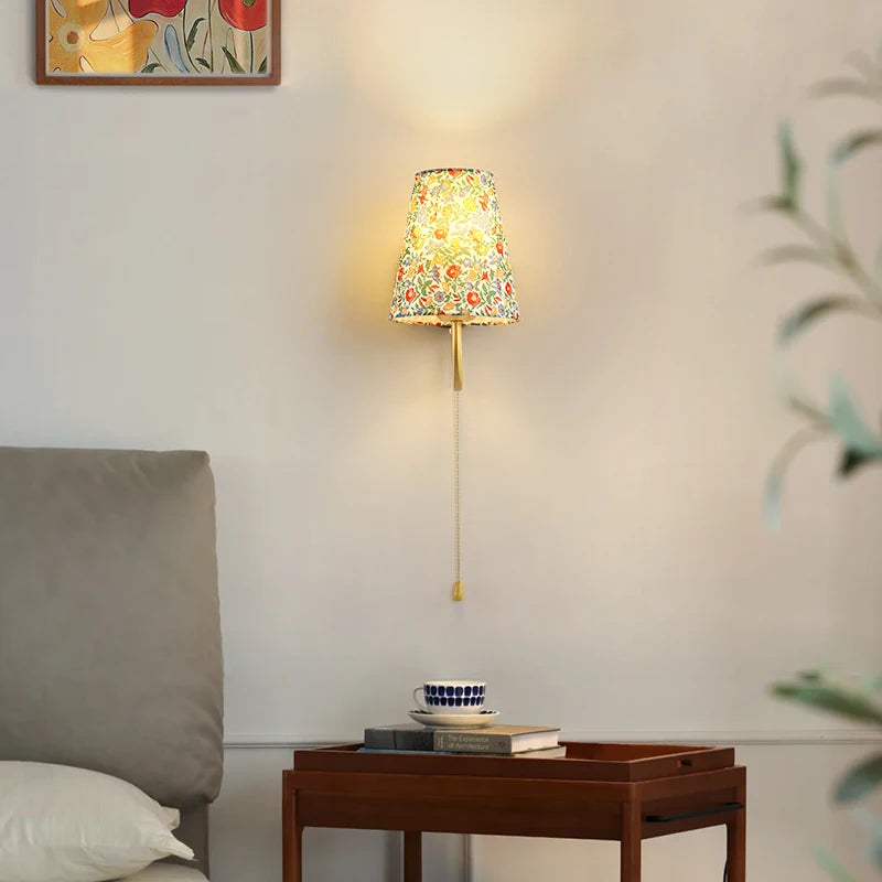 Decorative wall lamp with colourful shade on a wall next to a wooden side table with books and a cup. Modern wood and fabric bedside wall sconce with E27 base.