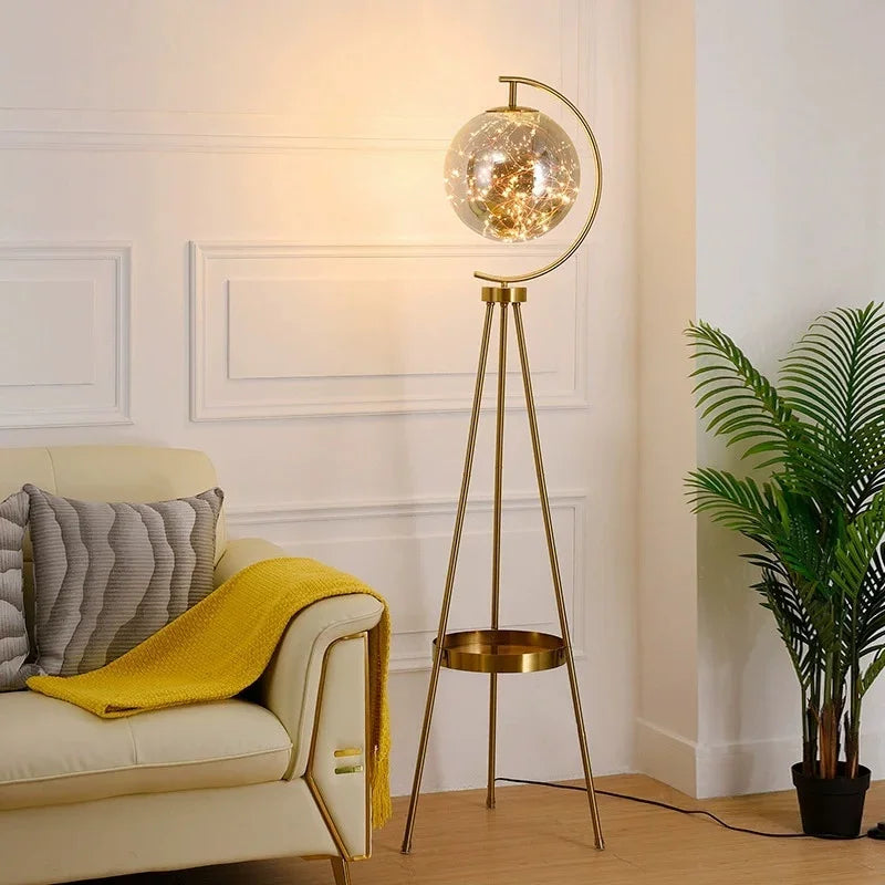 Gold floor lamp with a glass shade in a living room setting. Nordic tripod glass floor lamp with gold finish for a modern living room.