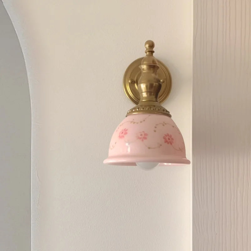 Wall-mounted light fixture with pink glass shade and gold metal fixture on a white wall.